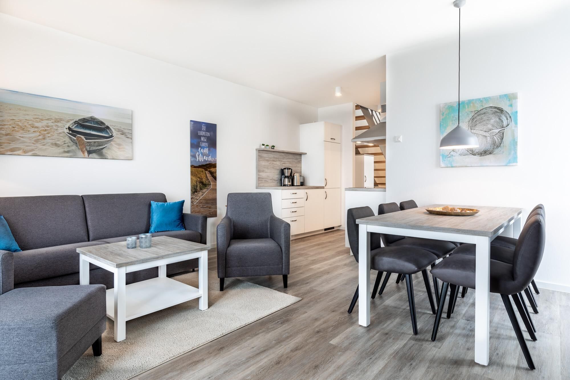 Ein modernes Wohnzimmer mit einer Couch, Sesseln, einem Couchtisch und einem Essbereich mit Stühlen und einem Tisch. Im Hintergrund befindet sich eine Küche mit Herd und Schränken.