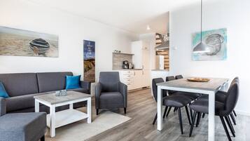 Ein modernes Wohnzimmer mit einer Couch, Sesseln, einem Couchtisch und einem Essbereich mit Stühlen und einem Tisch. Im Hintergrund befindet sich eine Küche mit Herd und Schränken.