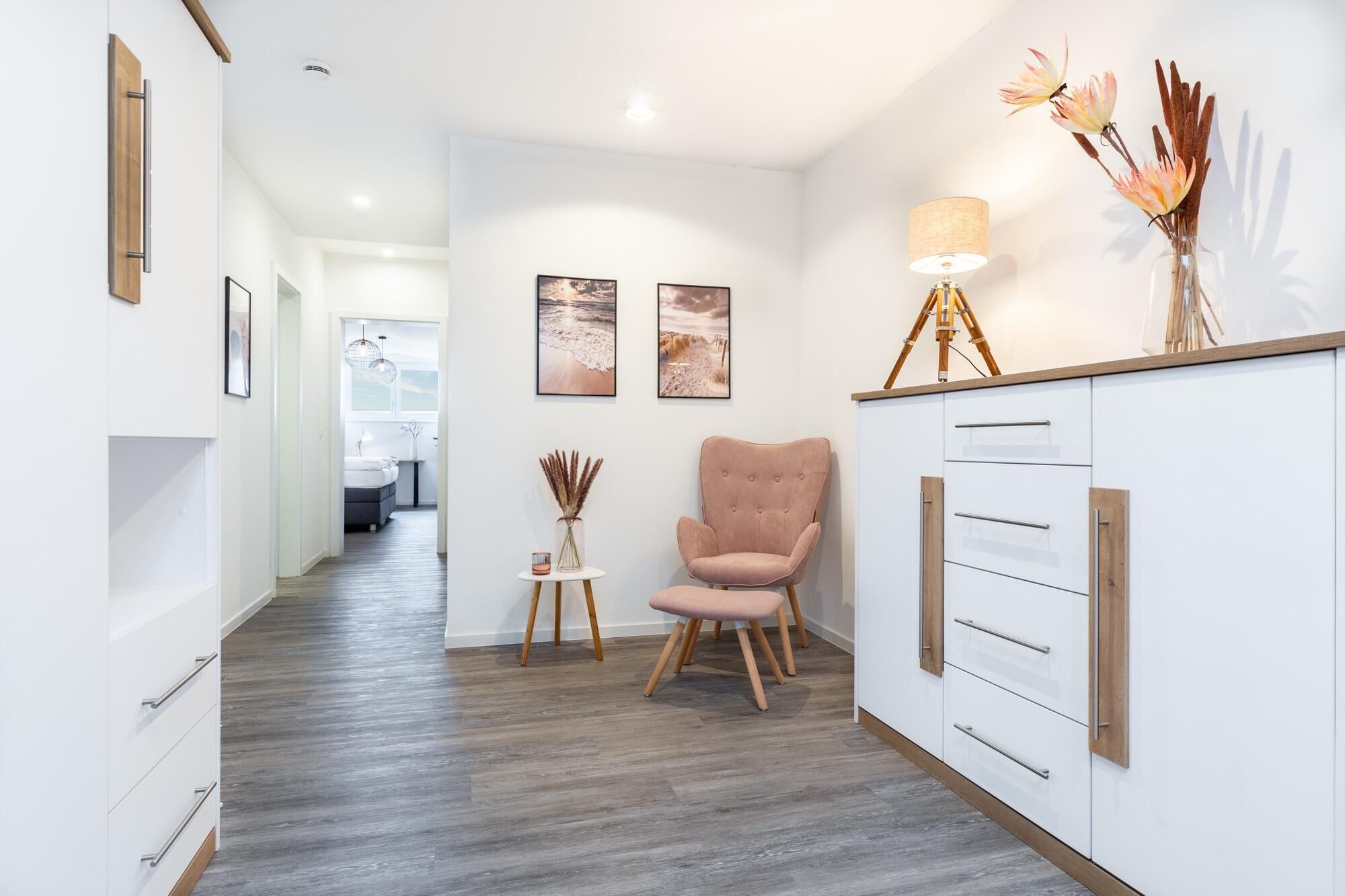 Un couloir moderne avec un plancher en bois, un fauteuil rose, une petite table avec une lampe et un meuble avec des tiroirs.