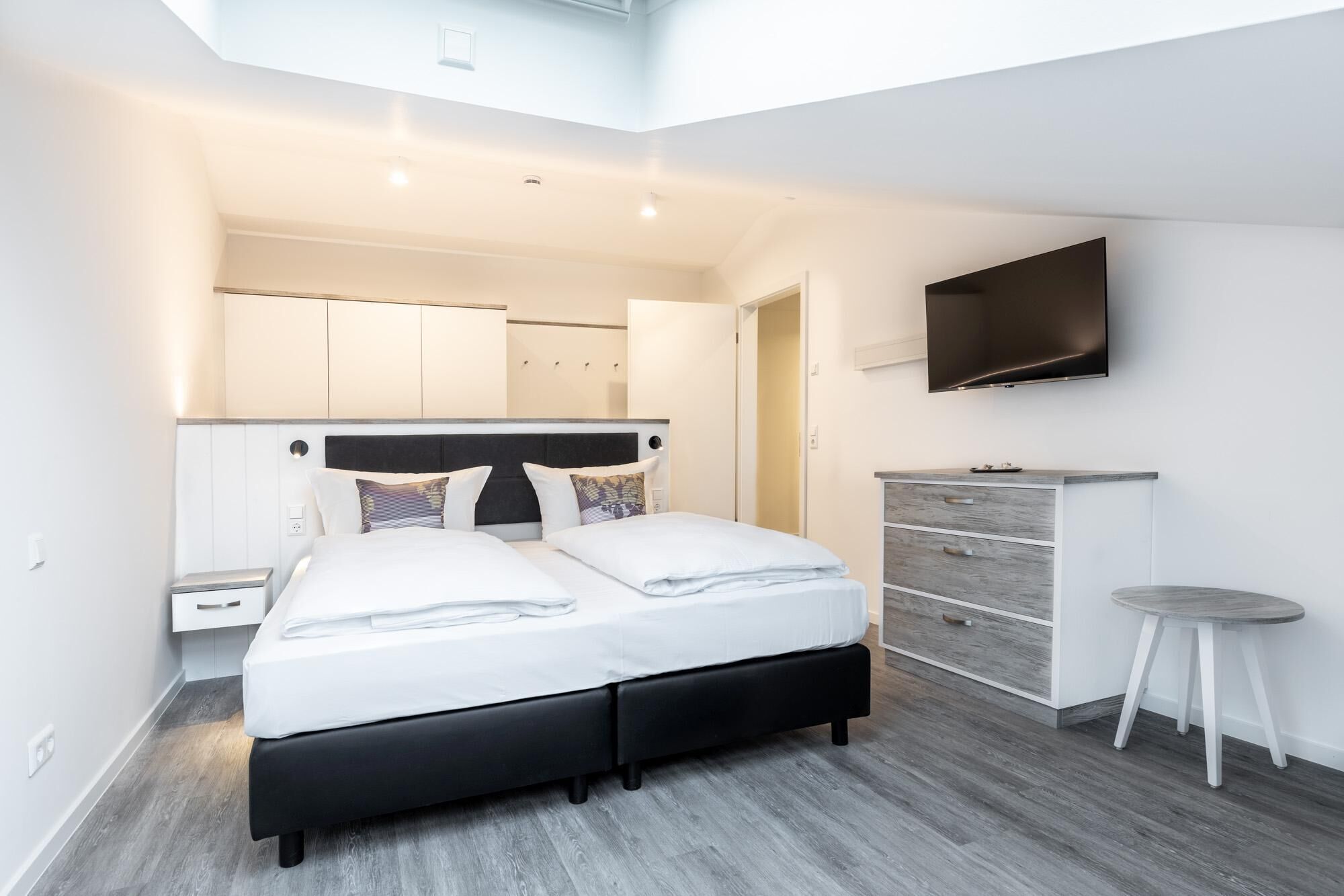 A modern bedroom with a bed, a desk, a TV, and a small stool.