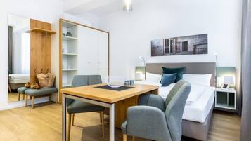 Une chambre d’hôtel moderne avec une table à manger en bois, deux chaises grises à revêtement en tissu, un lit avec une literie blanche et un oreiller vert, une table de chevet avec une lampe, et un grand miroir au mur.