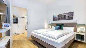 Une chambre à coucher moderne avec un lit, des tables de chevet, une commode, une œuvre d’art accrochée au mur et un espace cuisine visible.