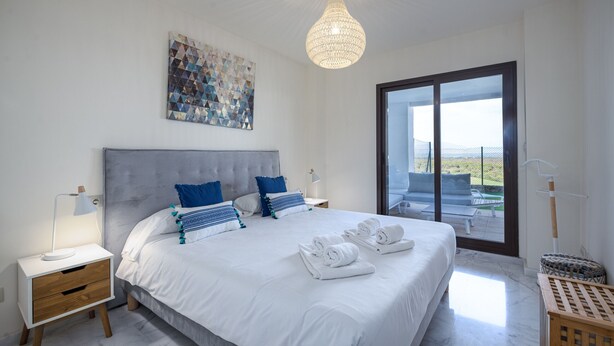 A modern bedroom with a large bed, a headboard, a nightstand, a lamp, a painting, and a view through a glass door. - Alcaidesa Golf and Beach 2356 (La Alcaidesa)