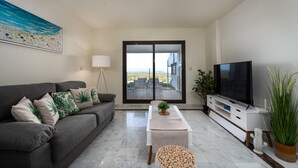 A modern living room with a grey sofa, a flat-screen TV, a coffee table, and a view through large sliding doors to an outdoor patio. - Alcaidesa Golf and Beach 2356 (La Alcaidesa)