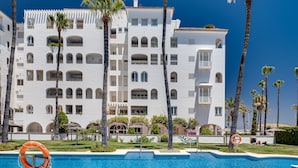 A swimming pool with a lifebuoy, surrounded by palm trees and a white building with multiple balconies.