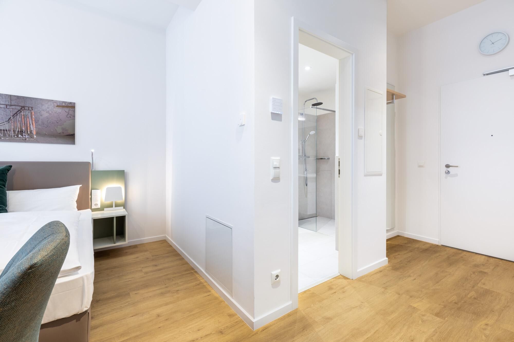 Moderni hotellihuone, jossa on puulattia, valkoisilla lakanoiden peitetty sänky, yöpöytä lampun kanssa ja lasinen suihkukaappi.