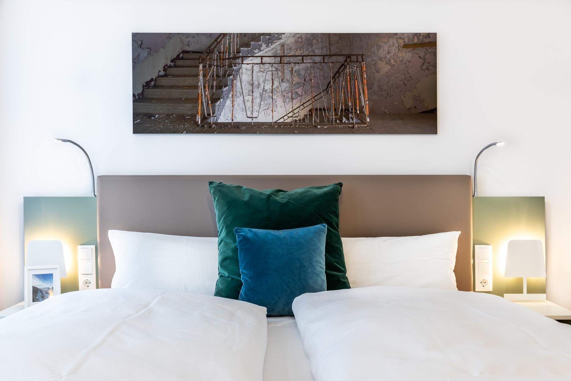 A hotel room with a bed, two bedside lamps, and a painting of a staircase.