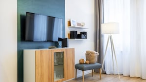 A modern living room with a flat-screen TV mounted on the wall, a wooden cabinet with glass doors, a floor lamp, and a small table with a basket.