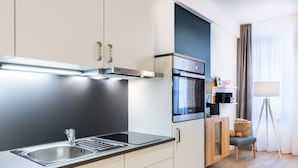 A modern kitchen with stainless steel appliances, a built-in oven, and a sink. There is a small seating area with a chair and a lamp.