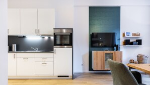 A modern kitchen with white cabinets, a built-in oven, and a microwave. There is a dining area with a wooden table and a green chair. A flat-screen TV is mounted on the wall above the cabinets.