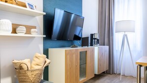 Una habitación de hotel moderna con una televisión de pantalla plana montada en la pared, un mueble de madera con puerta de vidrio, una cesta de mimbre con toallas y una lámpara de pie.