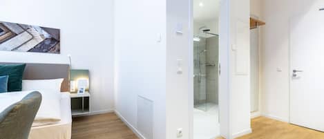 A modern hotel room with a wooden floor, a bed with a headboard, a bedside table with a lamp, and a glass shower enclosure.