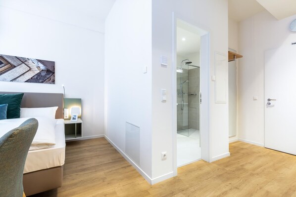 Una habitación de hotel moderna con suelo de madera, una cama con cabecero, una mesita de noche con lámpara y un box de ducha de vidrio.