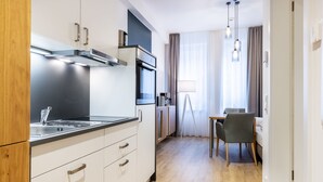 A modern kitchen with white cabinetry, a stainless steel range hood, and a built-in oven. There is a dining area with a table and chairs, and a window with curtains.