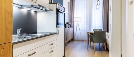 Een moderne keuken met witte kasten, een roestvrijstalen afzuigkap en een ingebouwde oven. Er is een eethoek met een tafel en stoelen, en een raam met gordijnen.