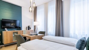 A hotel room with a bed, a desk with a lamp, a TV, and a large window with curtains.