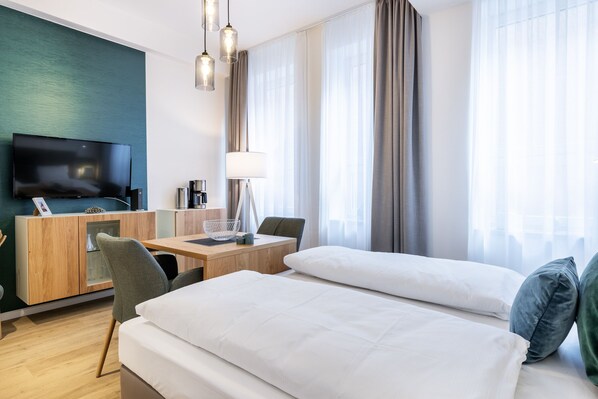 Una habitación de hotel con una cama, un escritorio con lámpara, un televisor y un ventanal con cortinas.