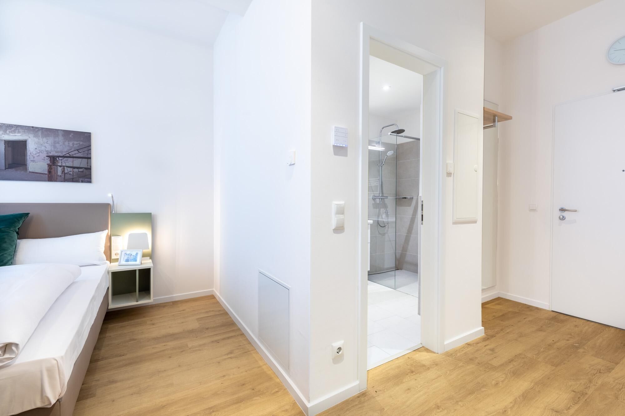 Une chambre d’hôtel moderne avec un plancher en bois, un lit avec du linge de maison blanc, une table de chevet avec une lampe et un espace douche.