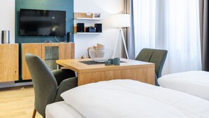 A hotel room with a flat-screen TV, a wooden desk, two chairs, a bed, a lamp, and a window with curtains.