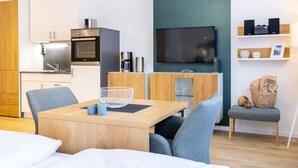 A modern hotel room with a wooden dining table, grey upholstered chairs, a TV mounted on a teal wall, and a kitchenette with a stainless steel refrigerator and oven. - Modernes, Hundefreundliches Apartment in Außergewöhnlicher Hafenlage von Wismar (Hansestadt Wismar)