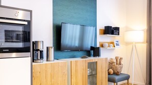 A modern kitchen with a built-in oven, a coffee maker, and a TV mounted on a teal wall. There is a wooden cabinet with glass doors and a small bench. - Modernes, Hundefreundliches Apartment in Außergewöhnlicher Hafenlage von Wismar (Hansestadt Wismar)