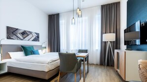 Habitación de hotel moderna con cama, escritorio, silla, lámpara, televisor y un cuadro en la pared.