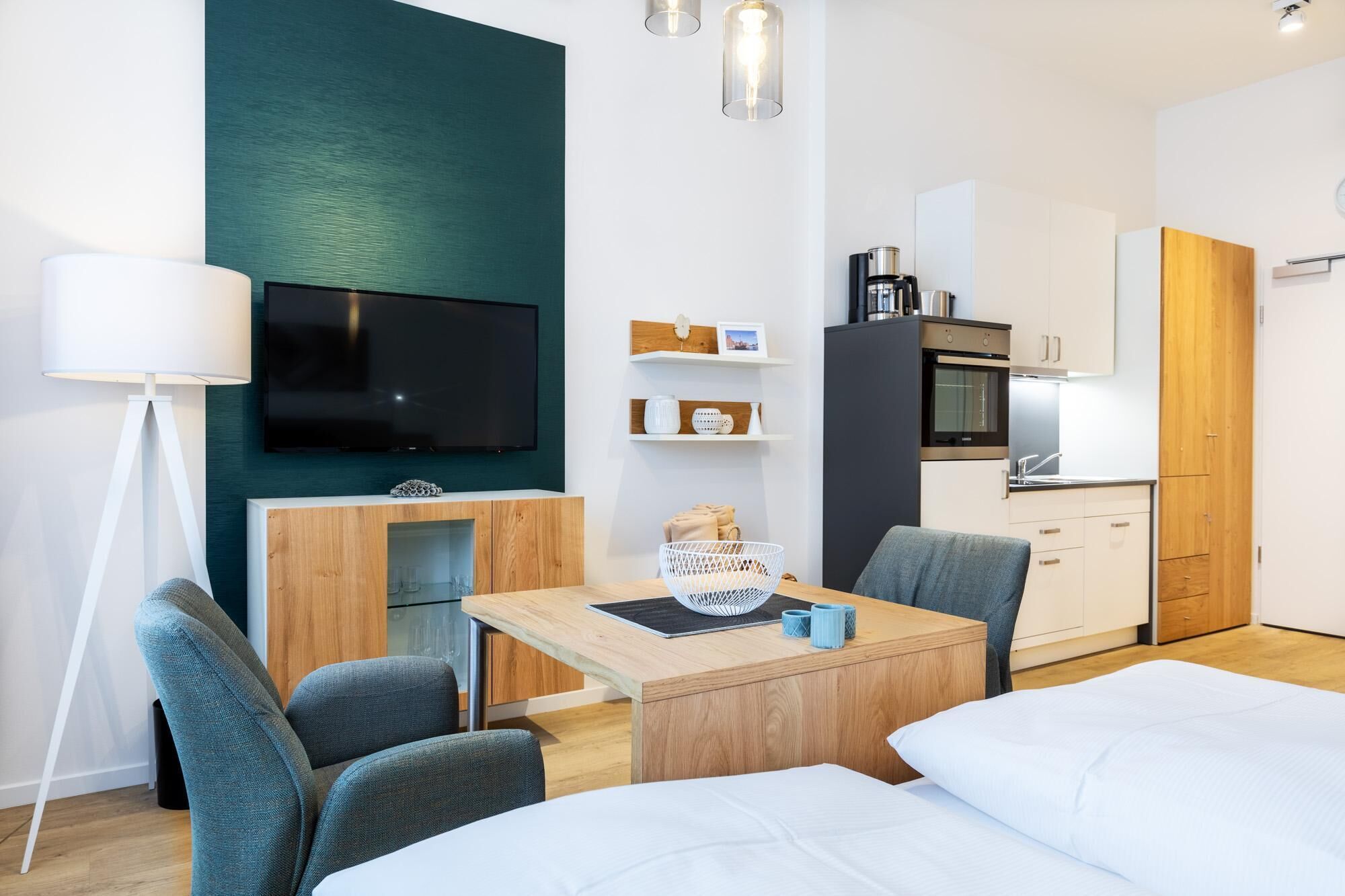 A modern hotel room with a TV, a dining table, two chairs, a bed, a kitchenette, and a large window.