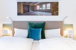 Quarto de hotel com uma cama grande, duas luminárias de cabeceira e um quadro na parede retratando um espaço interior.