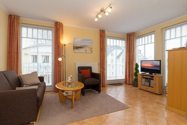 Una sala de estar con sofá, sillón, mesa de centro, mueble para televisión y ventanas amplias.