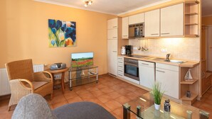 A modern kitchen with white cabinets, a microwave, and a coffee maker. There is a dining area with a glass table and chairs. A painting is on the wall, and a television is on a stand.