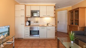 A compact kitchen with a microwave, oven, and sink, adjacent to a living area with a glass coffee table and a plant.