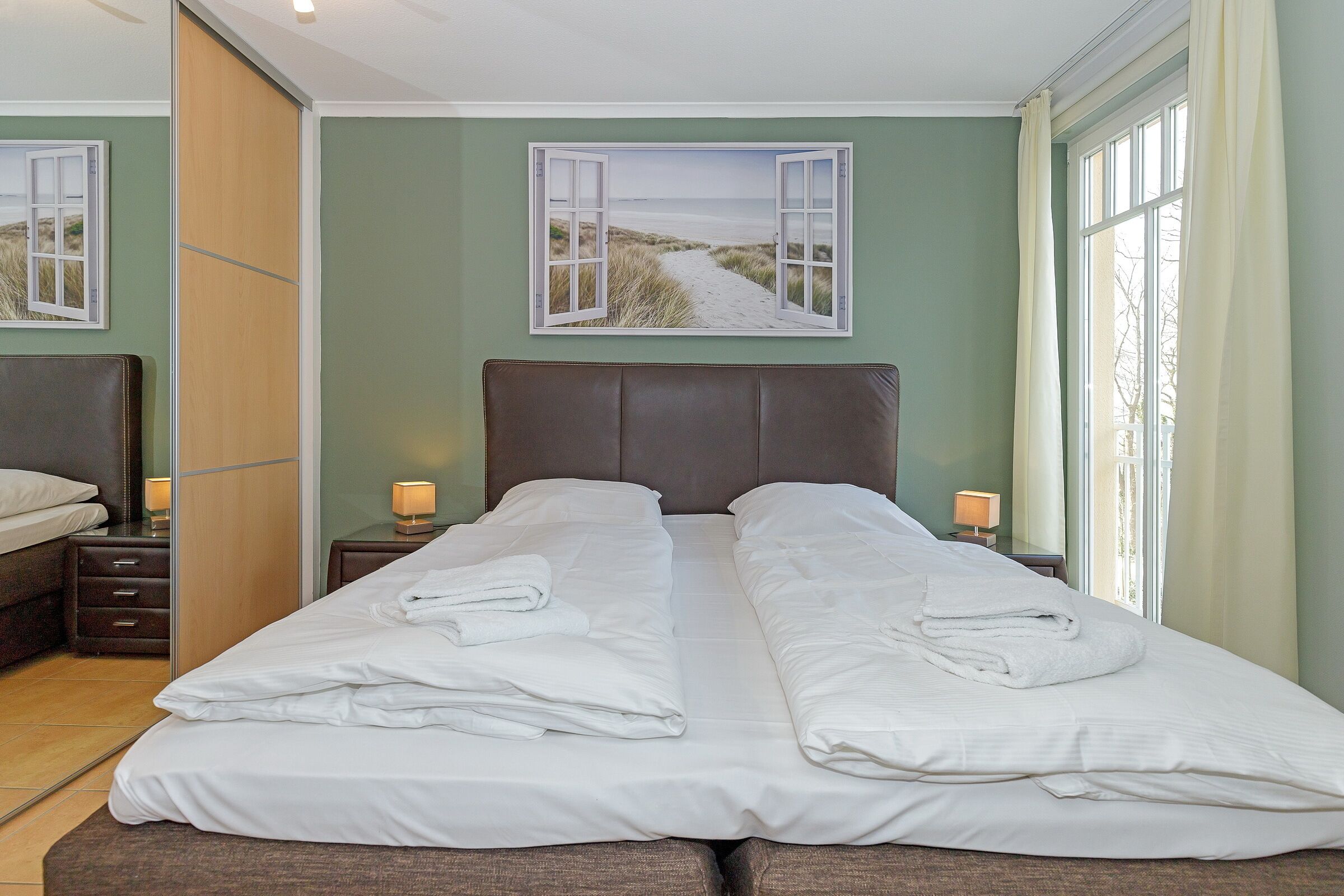 Een tweepersoonsbed met witte lakens en handdoeken, een houten hoofdbord, nachtkastjes met lampen, en een groot raam met gordijnen.