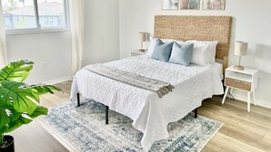 Une chambre à coucher avec un lit, une table de chevet, une lampe, une grande plante et une fenêtre avec des rideaux.