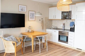 A modern kitchen with white cabinets, a built-in oven, a microwave, and a sink. There is a dining table with chairs and a wicker chair with cushions.