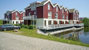A row of red and white houses with balconies, a paved walkway, and a body of water. - Great vacation home on the dyke with terrace, balcony with sea view, sauna & wood-burning stove (Nordseeheilbad Schillig)