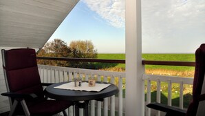 A balcony with a white railing, a table with two chairs, and a view of a green field and trees. - Great vacation home on the dyke with terrace, balcony with sea view, sauna & wood-burning stove (Nordseeheilbad Schillig)