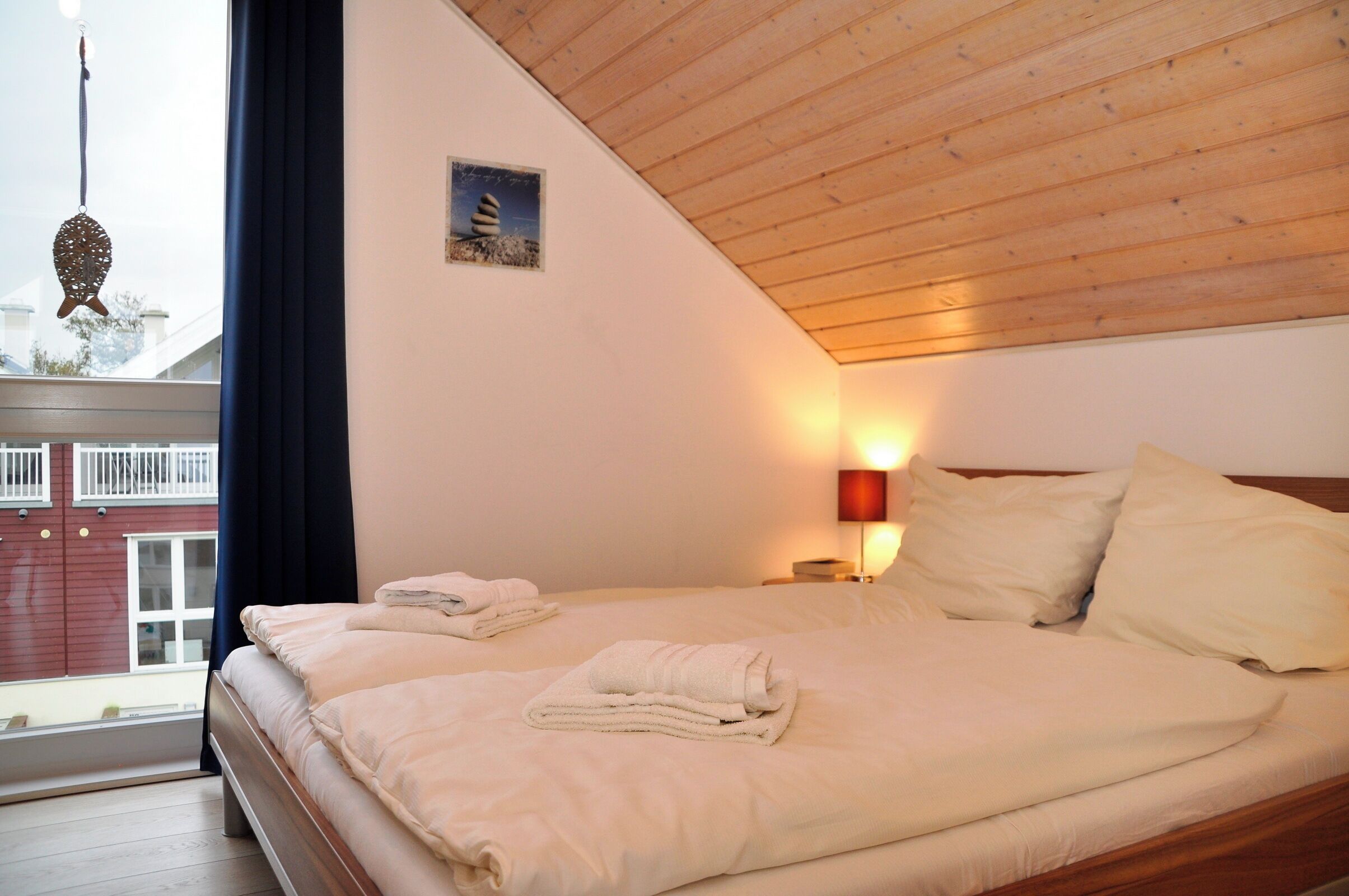 A bedroom with a wooden ceiling, two beds with white bedding, a wall-mounted lamp, and a view of a building outside.