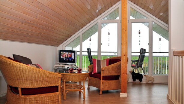 A cozy living room with wicker furniture, a television, and a balcony with a view. - Great vacation home on the dyke with terrace, balcony with sea view, sauna & wood-burning stove (Nordseeheilbad Schillig)