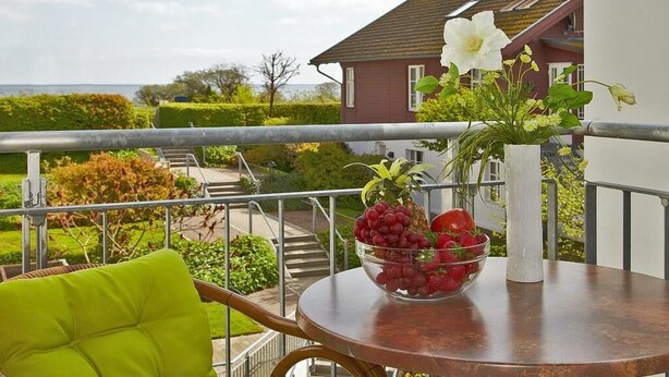 A balcony with a table holding a bowl of fruit and a vase with flowers, overlooking a garden and a house.