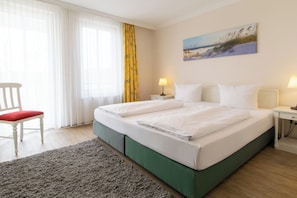 A neatly made double bed with white linens, a green upholstered headboard, a small bedside table with a lamp, a chair with a red cushion, and a painting of a beach scene on the wall.