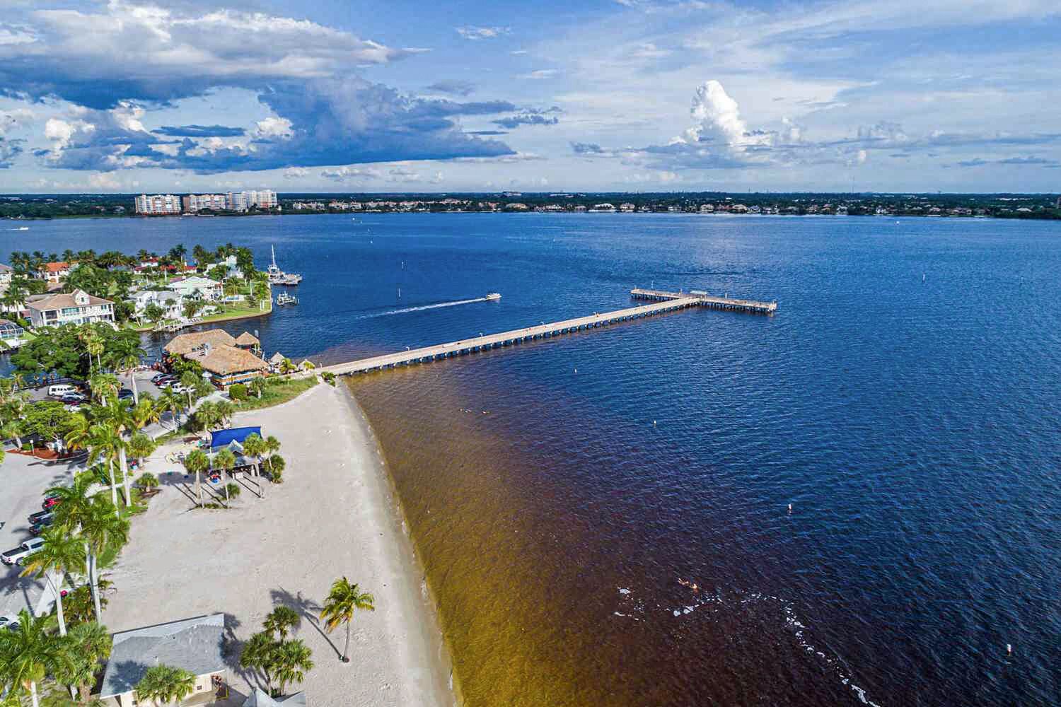 Une zone côtière avec une longue jetée qui s’avance dans l’eau, une plage bordée de palmiers, et en arrière-plan, des bâtiments et le profil d’une ville lointaine.