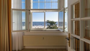 Une pièce dotée d’une grande baie vitrée donnant sur une plage, d’un radiateur placé sous la fenêtre et d’un plancher en bois.