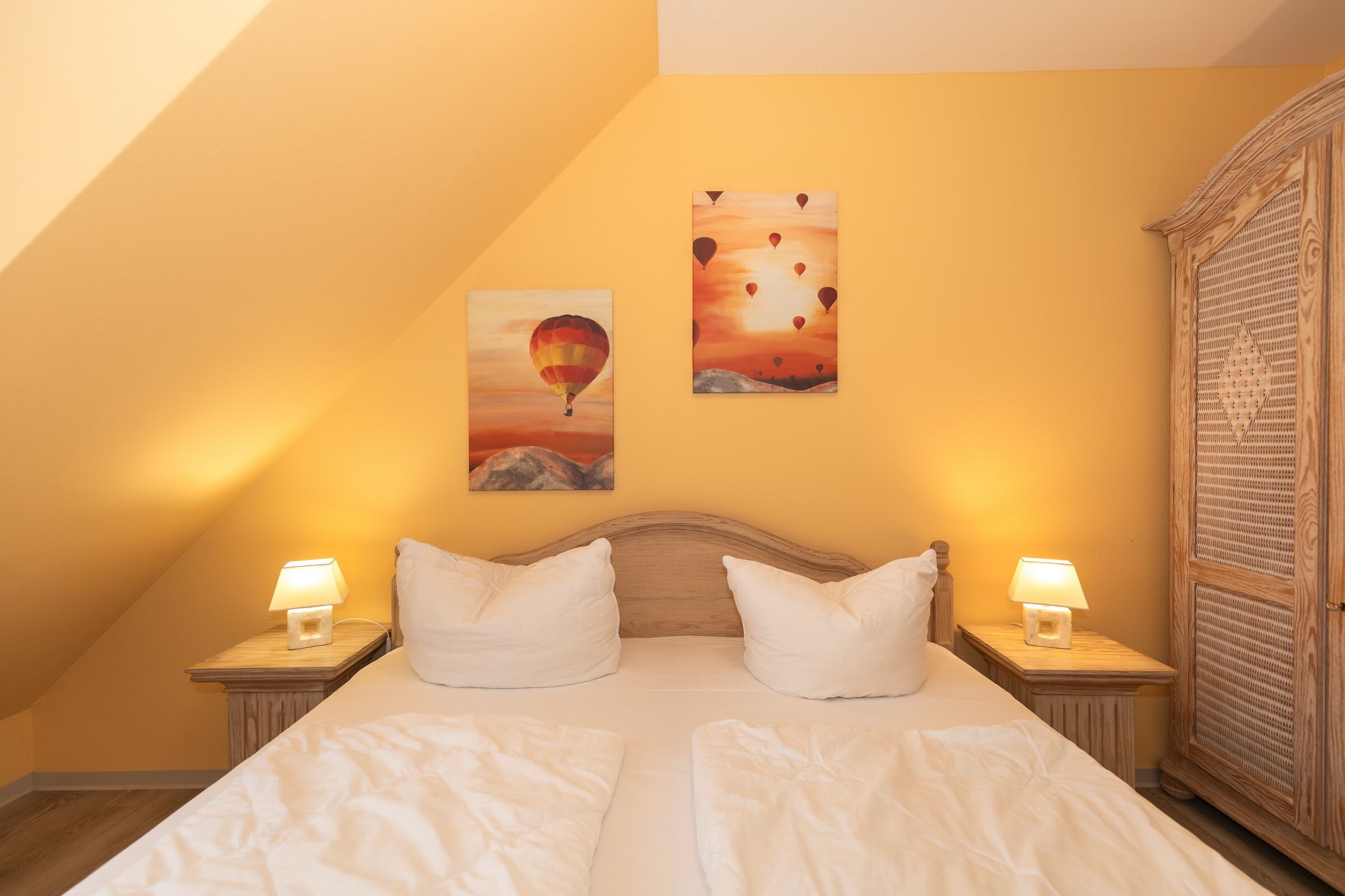 Een slaapkamer met een schuine kap, twee bedlampen, een bed met witte lakens en twee schilderijen aan de muur.