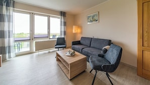 A modern living room with a sofa, armchair, and coffee table, large windows with curtains, and a painting on the wall.