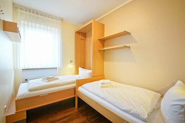 A room with two single beds, wooden headboards, a window with sheer curtains, and a built-in shelf above one bed. - Strandnahe, Lichtdurchflutete Ferienwohnung an der Nordsee mit Meerblick u. Wlan (Nordseeheilbad Schillig)