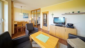 A living room with a wooden coffee table, a TV, and a sofa. There is a dining area with a table and chairs. - Strandnahe, Lichtdurchflutete Ferienwohnung an der Nordsee mit Meerblick u. Wlan (Nordseeheilbad Schillig)