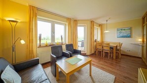 A living room with a sofa, two armchairs, a wooden coffee table, a dining table with chairs, and a painting on the wall. - Strandnahe, Lichtdurchflutete Ferienwohnung an der Nordsee mit Meerblick u. Wlan (Nordseeheilbad Schillig)