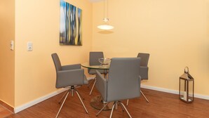 A dining area with a glass table, four grey chairs, a hanging light, and a painting on the wall.