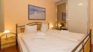 A double bed with white linens, a bedside table with a lamp, a painting on the wall, and a mirror reflecting the room.