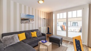 Ein modernes Wohnzimmer mit grauem Sofa, Hocker und Streifen-Tapete. Ein Strandbild hängt an der Wand, und es gibt einen Blick auf den Balkon.
